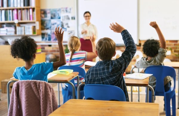 Children in a classroom, raising their hands; a teacher at the front of class.