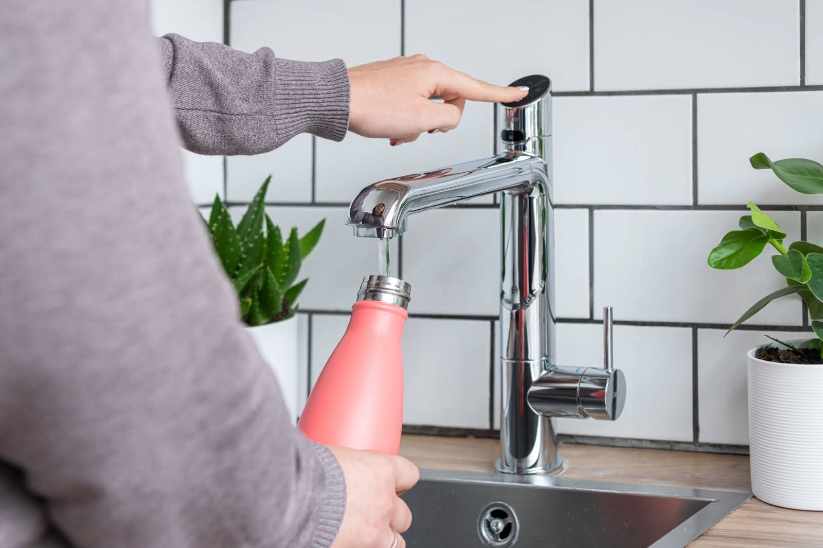 A person filling up a bottle from a silver HydroTap, with two plants next to it.