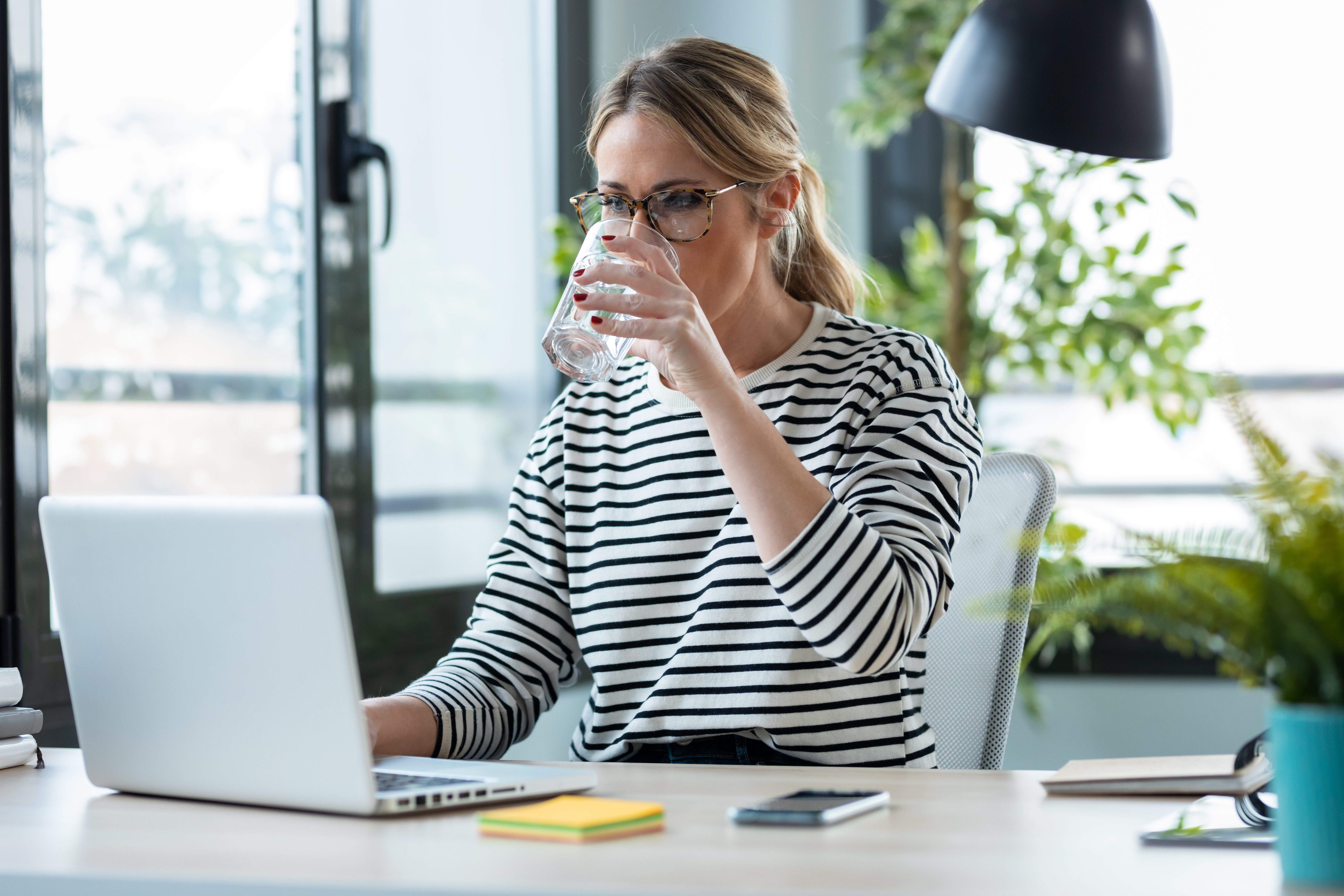 A woman working on a laptop drinking water at a desk