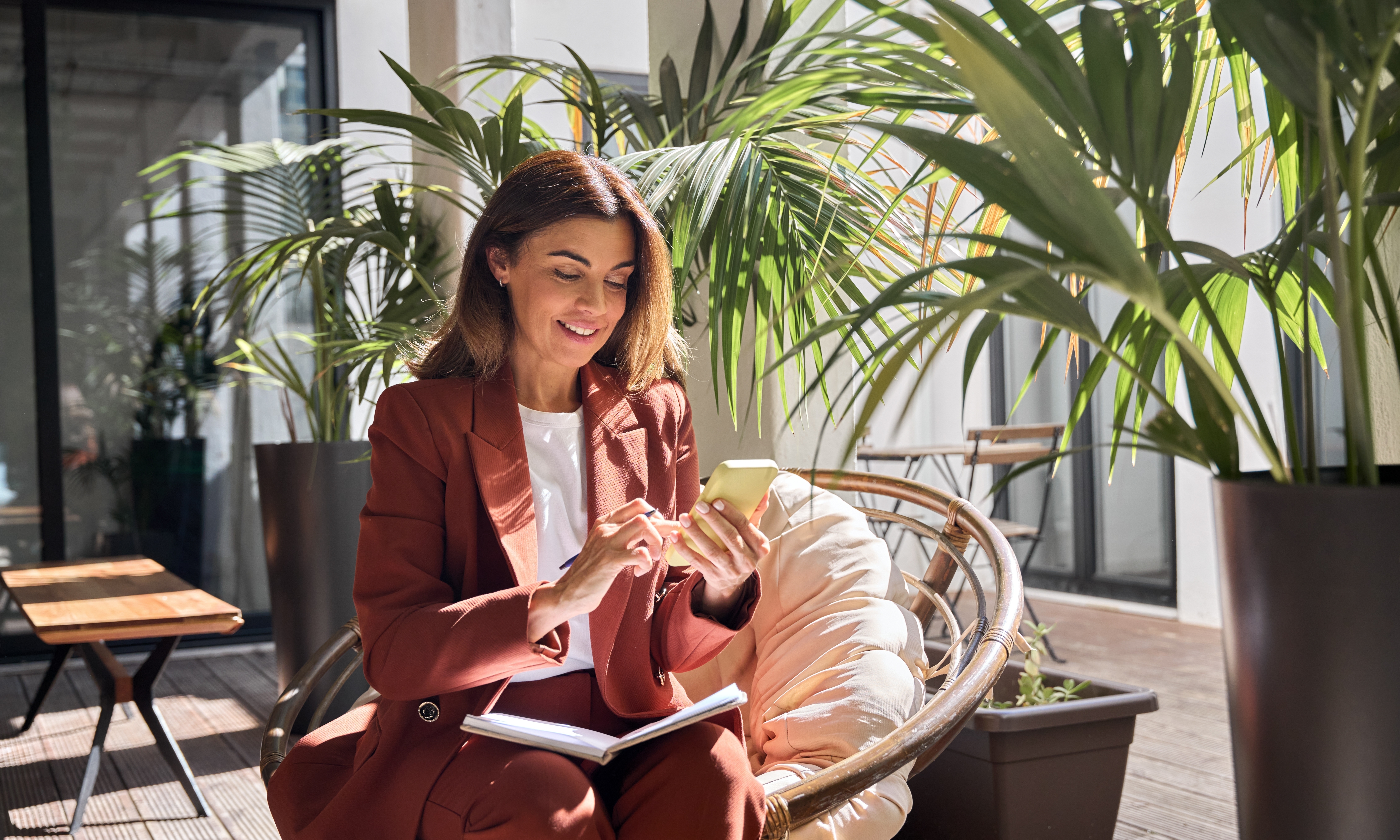 A woman using her phone with an open notebook, surrounded by indoor plants